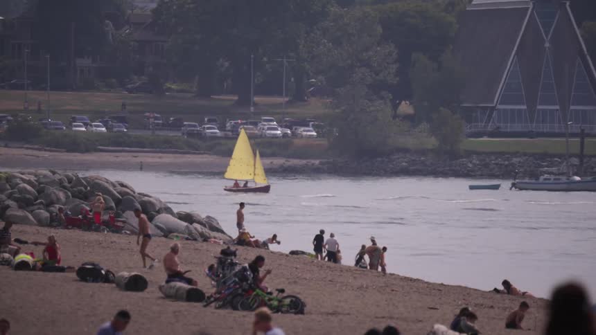 The video shows a busy beach scene on a sunny day in the background a sailboat with a bright yellow sail glides across the water