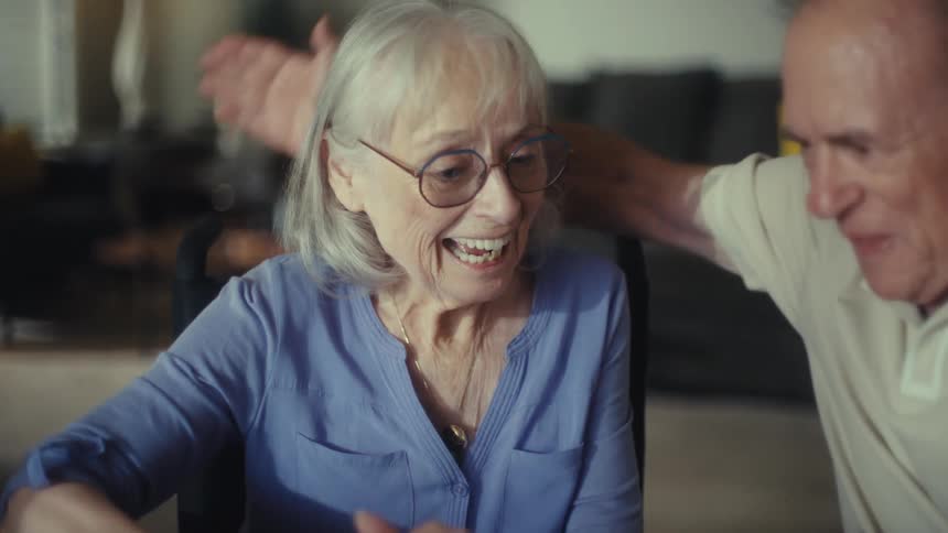 The video shows an older couple sitting at a table having a conversation the woman is wearing a blue shirt and glasses and the man is wearing a white collared shirt