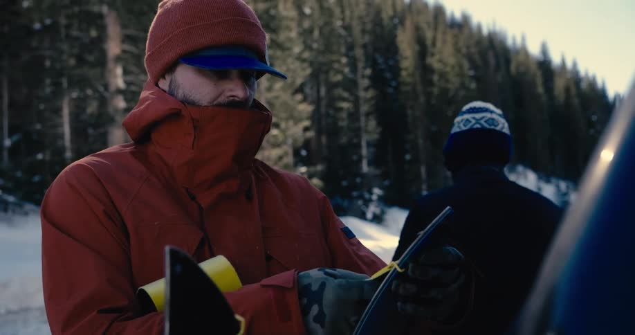The video shows a closeup shot of two people preparing to ski or snowboard the person in front is wearing a red jacket and hat and the person in the background has on a patterned beanie