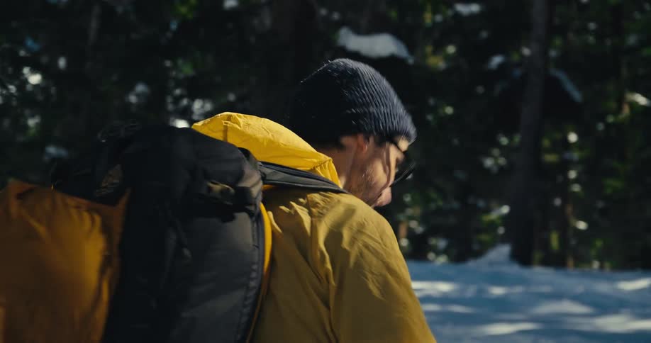 A person wearing a yellow jacket and a backpack walks through a snowy landscape turning to look at the camera and then continuing on their way using ski poles for movement