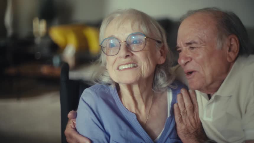 An older couple with gray hair is sitting close together and smiling the woman wearing a blue shirt and glasses is in a wheelchair the man wearing a creamcolored shirt is leaning in and has his arm wrapped around her