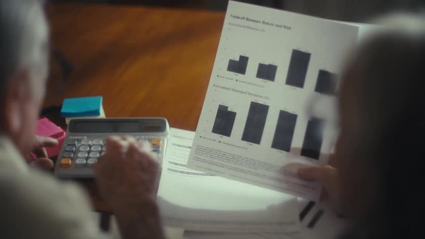 Two elderly people analyze printed financial charts while using a calculator on the table