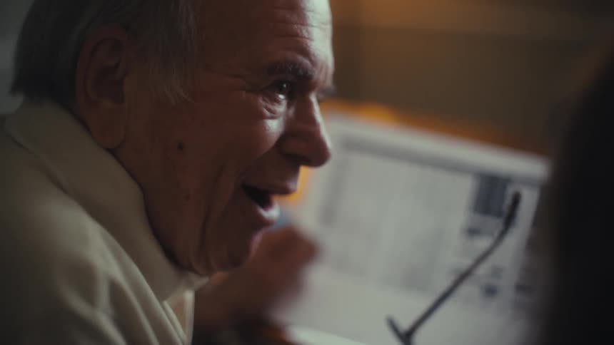 An older man is sitting at a table looking at some documents he is wearing a creamcolored shirt and he smiles as he takes off his glasses another person is sitting across from him