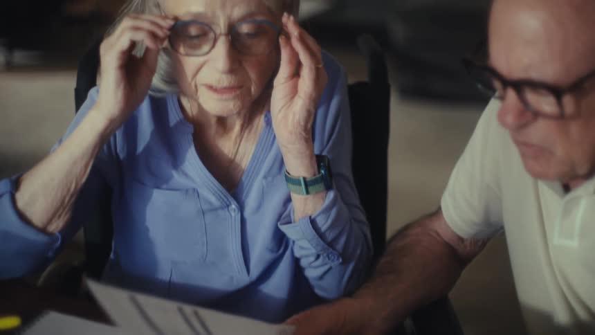 Two elderly sit at a desk with papers and a calculator and the woman is with a worried expression