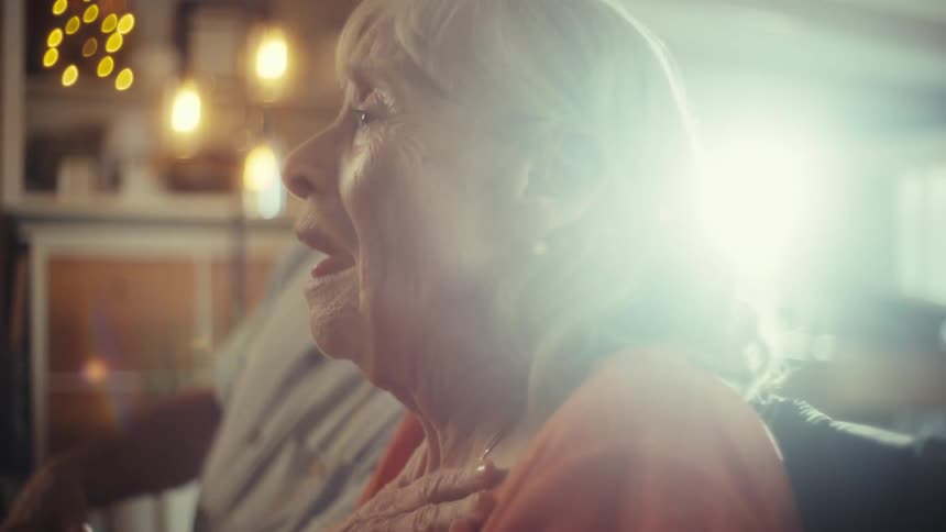 In a warmly lit room an elderly couple sings a heartfelt karaoke duet with the woman expressively placing her hand over her chest as they share a joyful musical moment