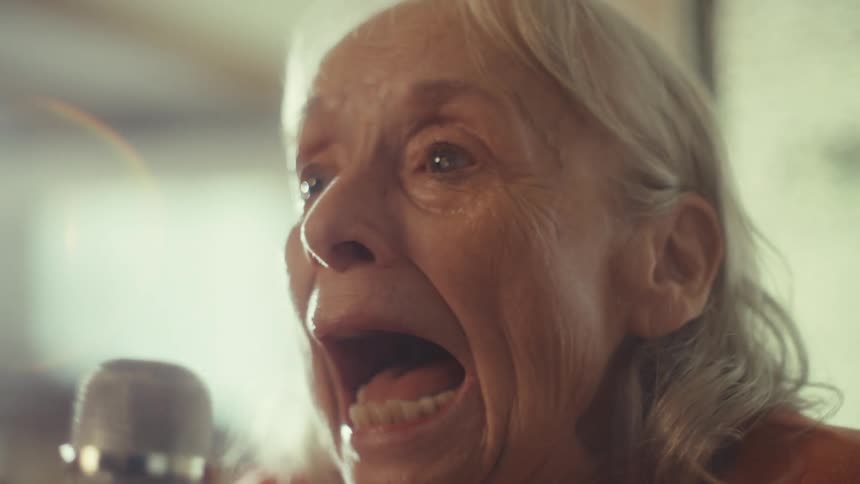 An elderly woman sings karaoke into a microphone indoors having a fun time at midday as sunlight reflects into the room