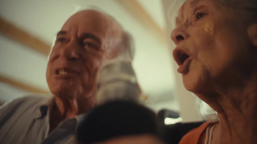 A close view shows a smiling elderly woman and a man both singing into a microphone with a bright light shining on their faces from the left