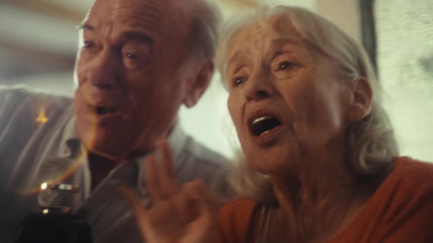 A close view shows a smiling elderly woman and a man both singing into a retro microphone with happy open mouthed expressions on their faces