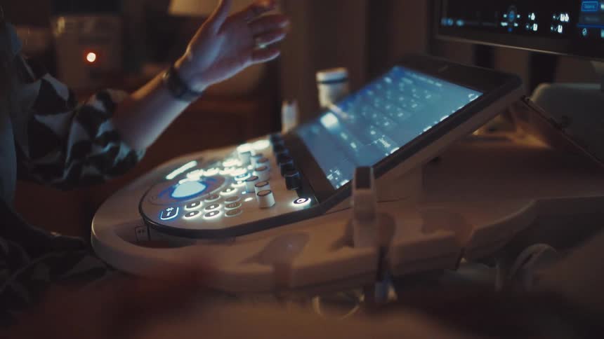 A doctor is operating an ultrasound machine a hand with a watch is shown pressing buttons and using the touchscreen on the console a large monitor in the background displays the ultrasound image