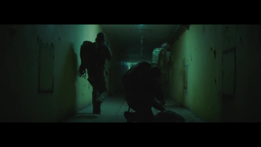 Several dark figures appearing to be soldiers move down a shadowy corridor their silhouettes are cast against the stark green walls of the hallway as they walk past a body lying on the floor