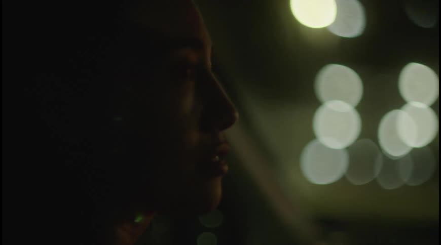 A darkskinned person is shown in profile sitting in a car at night the persons face is partially lit by the outoffocus city lights streaming in from outside