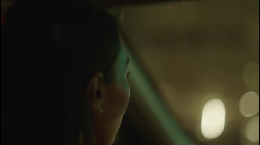 A darkhaired person is sitting in a car at night looking out the window the reflection of blurry colorful lights from outside streams across the window