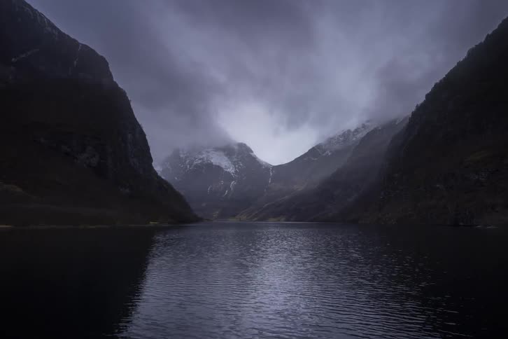 A dramatic shot shows a lake surrounded by mountains on both sides the sky is dark and stormy with gray clouds swirling around the mountaintops