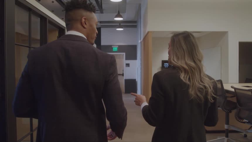 Two professionals walk through an office hallway while discussing work