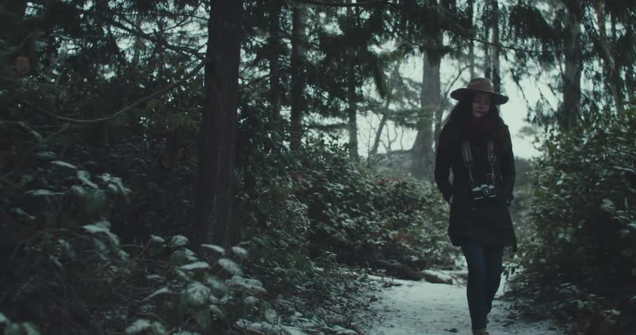 A woman wearing a long dark jacket a beige hat and a camera around her neck walks on a snowy path in a forest