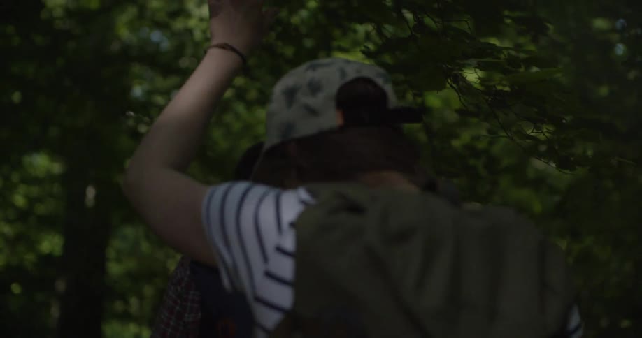 Two hikers with backpacks walk along a shaded forest path one in a striped shirt and hat the other ahead in a plaid shirt