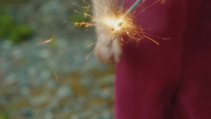 The provided video file shows a closeup of a person holding a lit sparkler the sparks are flying outward in all directions creating a bright energetic display the person is wearing a reddishpink garment the background is out of focus suggesting th