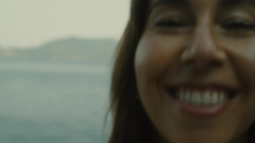 A smiling woman in a furlike wrap holds a bouquet of flowers while standing near the water with mountains in the distance