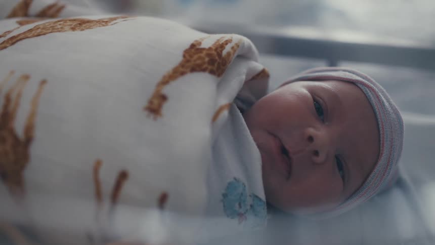 A shot focuses on a newborn baby wrapped in a swaddle with giraffe patterns lying peacefully in a bassinet the baby is wide awake looking around with a curious expression