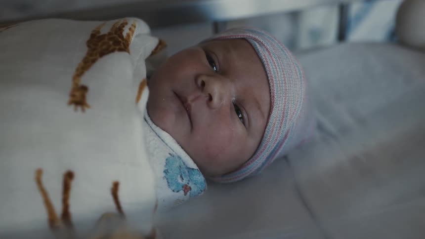 The first shot is a closeup of a newborn baby lying in a hospital bassinet the baby is wrapped in a blanket with a giraffe pattern and is wearing a striped beanie it is awake looking directly at the camera with wide eyes
