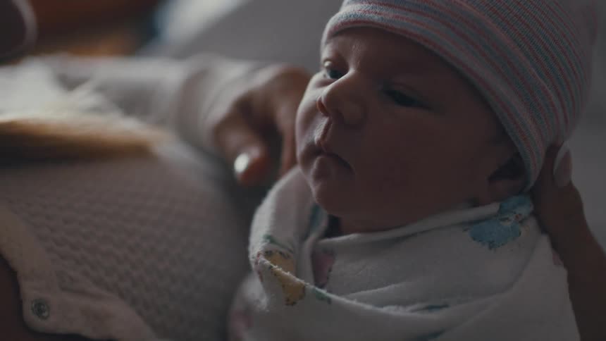 A shot focuses on a parent holding a newborn baby who is wrapped in a swaddle and wearing a hat the baby is held close to the parent with a profile view of the babys face