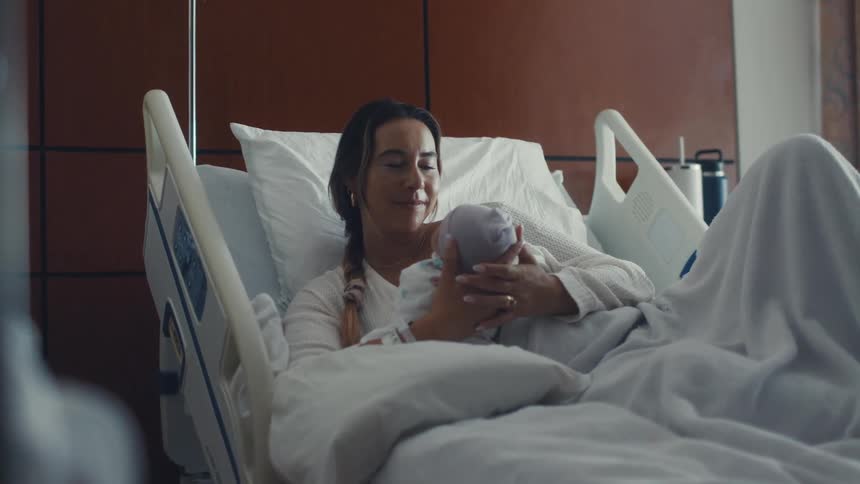 A smiling woman lies in a hospital bed gently holding a newborn baby wrapped in a blanket
