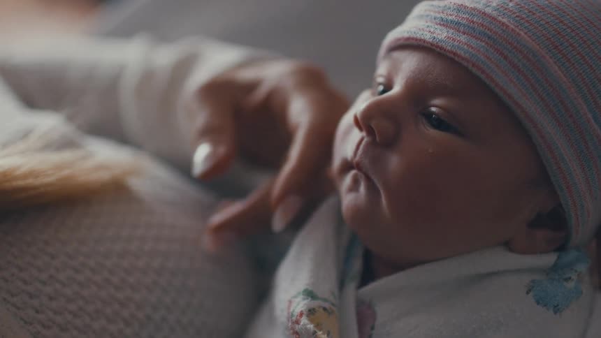 A newborn wrapped in a blanket and wearing a striped cap rests calmly while being gently held