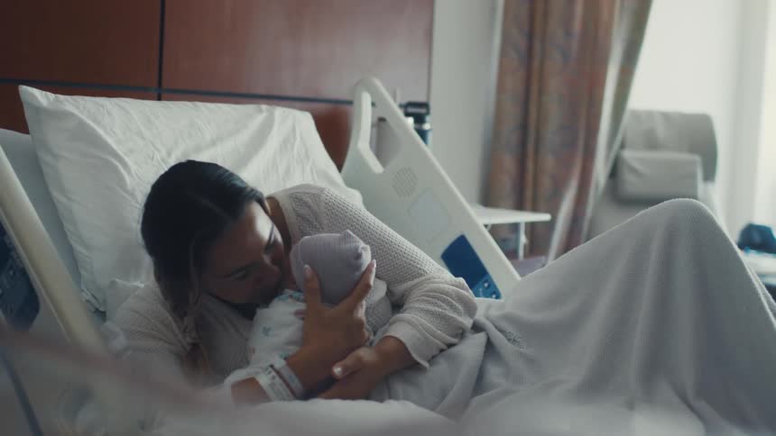 A new mother is seen lying in a hospital bed lovingly holding her newborn baby she gently brings the baby close to her face and gives it a tender kiss on its head