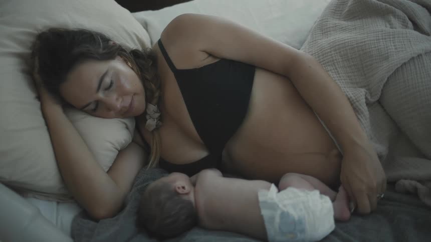 A tired new mother lies on her side in bed affectionately cradling her sleeping newborn baby she is resting her eyes closed as her baby peacefully sleeps by her side