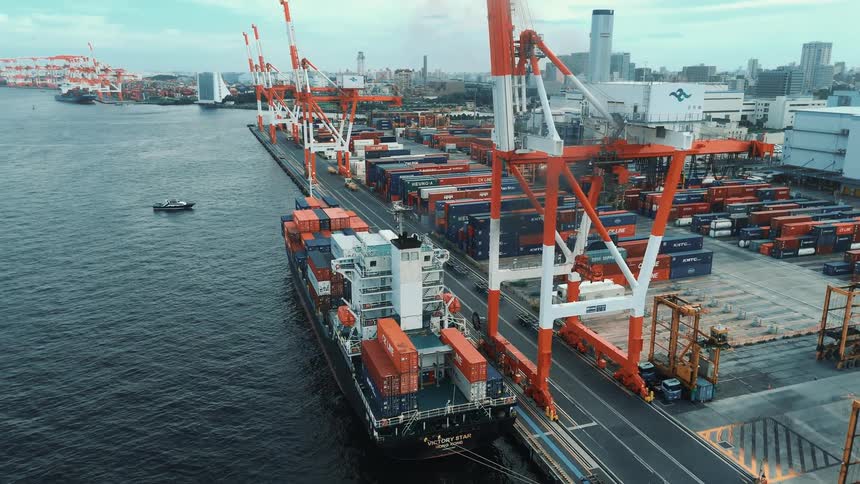A massive container ship is docked at a bustling industrial port with towering cranes lining the quay and a city skyline in the background