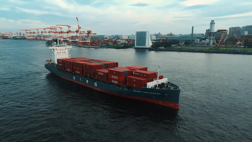 A cargo boat leaving a commercial port with a large shipping container depot
