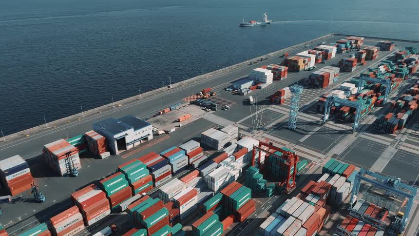 A boat approaching a commercial port with a large cargo shipping container depot while cargo trucks move around on the roads inside the depot