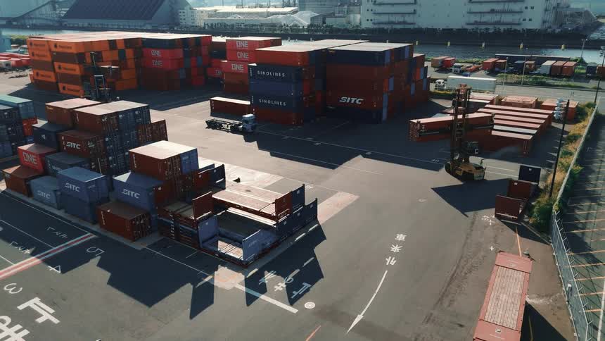 A seaport at midday alive with activity a forklift lifts and stacks cargo containers into towering rows while trucks move steadily in and out of the port along sunlit paved lanes transporting freight under the bright sky