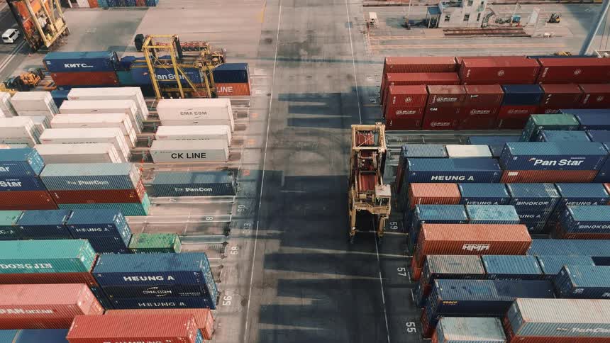 A loader carries a shipping container across a busy port at midday surrounded by towering stacks of colorful containers under the bright sun