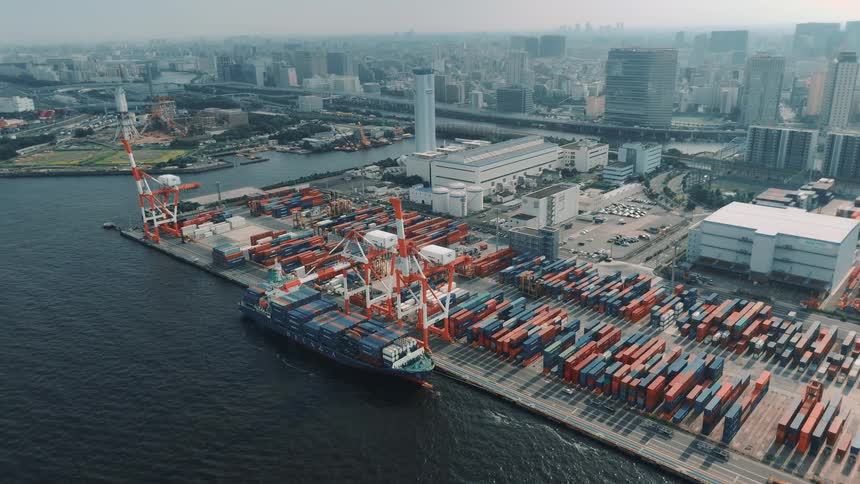 An aerial view of a seaport shows a cargo ship docked at the harbor with two towering cranes positioned above it the ships deck is stacked high with shipping containers while more containers are neatly piled across the port with the city behind
