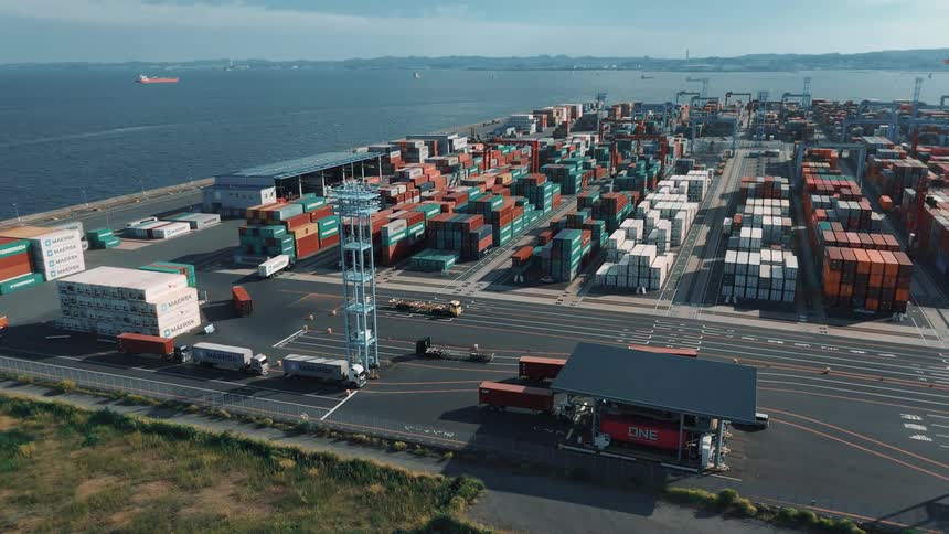 Rows of colorful shipping containers fill a busy port terminal beside the ocean with trucks and cranes in motion