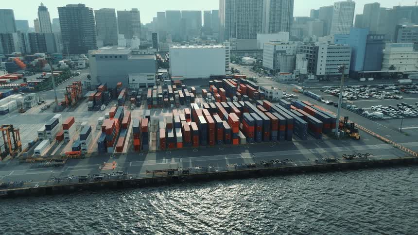 This drone footage captures an aerial view of a shipping port a large number of shipping containers are stacked on the dock with some trucks and other vehicles visible in the background a bustling city skyline provides a sharp contrast to