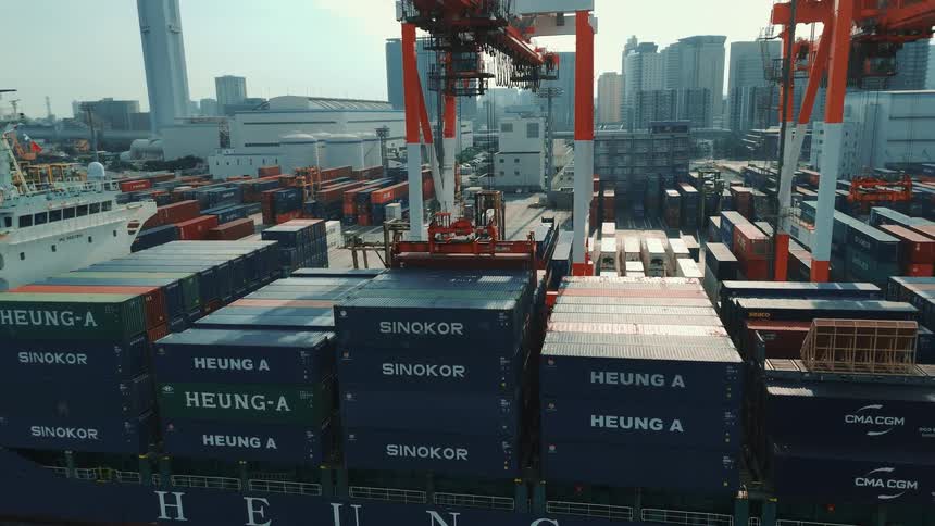An aerial shot shows a massive cargo ship docked at a port with many colorful shipping containers on it a large crane with a buildingsized arm moves a container and places it on the ship