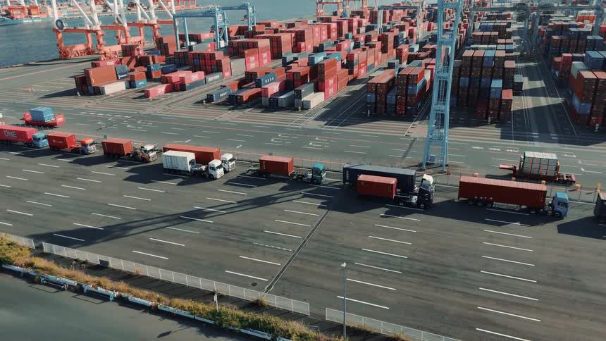 Trucks loaded with shipping containers drive out of a busy seaport at midday passing towering stacks of cargo containers under the bright sun