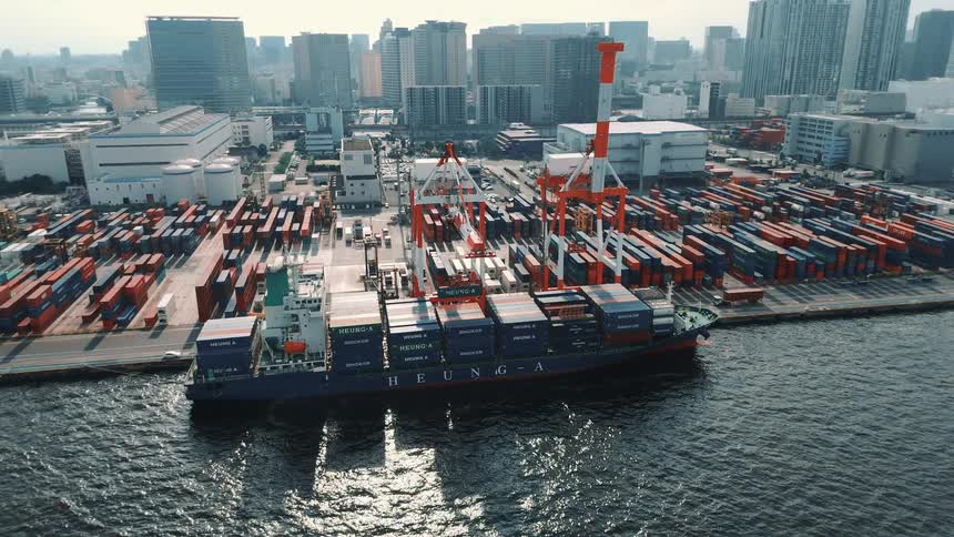 A cargo ship docked at a seaport stacked with containers with a crane positioned overhead and trucks moving along the port the city skyline rising in the background under the bright midday sun