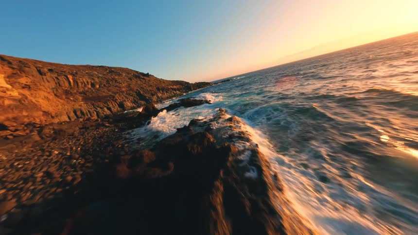 This is a fastpaced fpv first person view drone video of a rugged coastline the drone flies low and close to the rocky cliffs with waves crashing against the shore and the ocean reflecting the bright setting sun