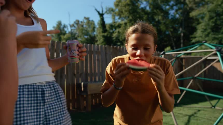 A smiling young girl in a brown tshirt is eating a large slice of watermelon while standing with other children and a woman in a sunny backyard