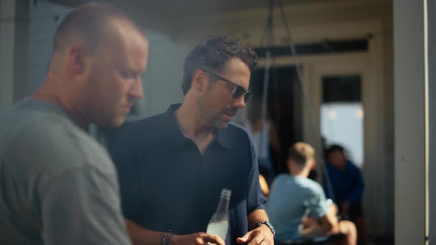 Two men are talking and drinking in the foreground while blurred figures of children and adults interact in the background on a patio