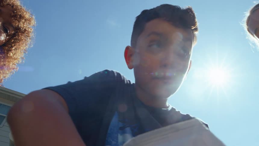 Four teenagers stand together on a sunny day each reaching into a shared bag of chips to grab a piece then eating it in the warm light