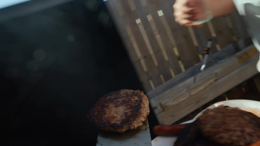 A man cooks burgers on a grill with a spatula while another man approaches with a beverage