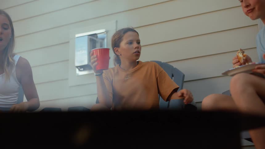 A young person in an orange shirt sits back in a chair outdoors sipping from a red plastic cup while relaxing with friends who are eating