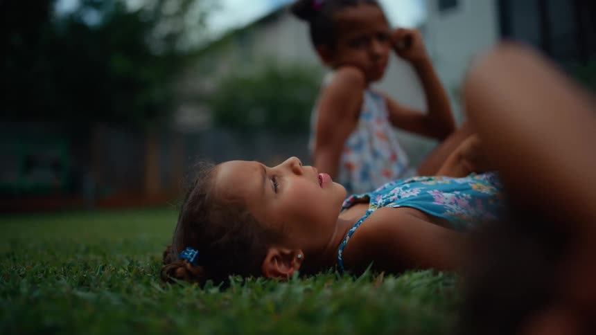 Two young girls are lying on the grass outdoors with one in the foreground looking up