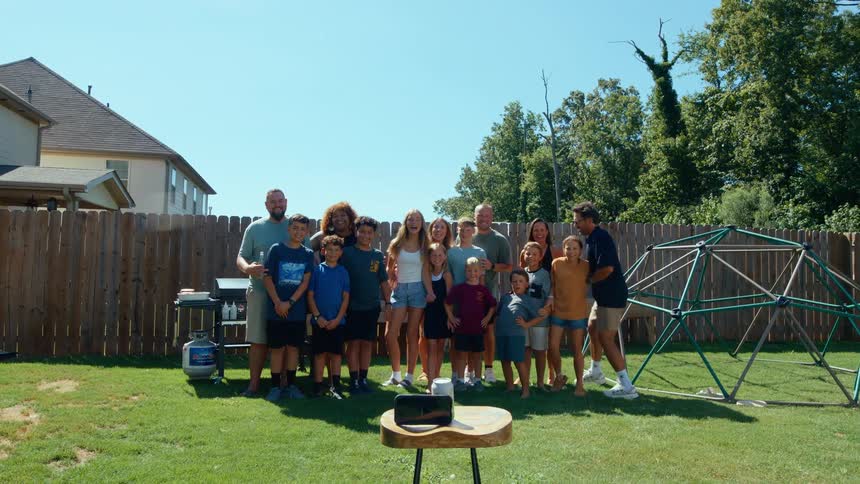 A large family group of men women and children of various ages gathers in a backyard with a geometric climbing frame as one man smiles and poses for a selftimer photograph on a smartphone