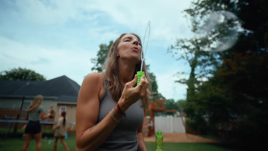 A woman stands in a sunny backyard at midday blowing shimmering bubbles from a bubble maker nearby another girl plays joyfully among the drifting bubbles while a few kids bounce on a trampoline in the background