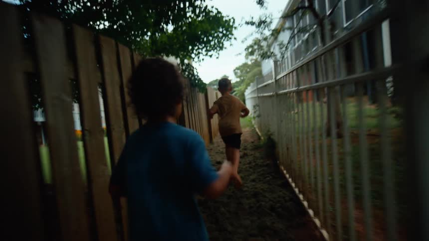 Two young boys run barefoot through a muddy path between a wooden fence and a metal fence
